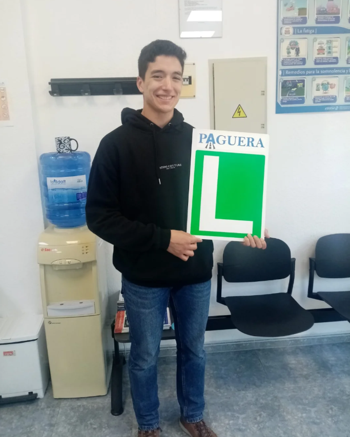 Joven sosteniendo cartel con letra 'L' grande y la palabra 'Paguera' en la parte superior en un aula de clases.