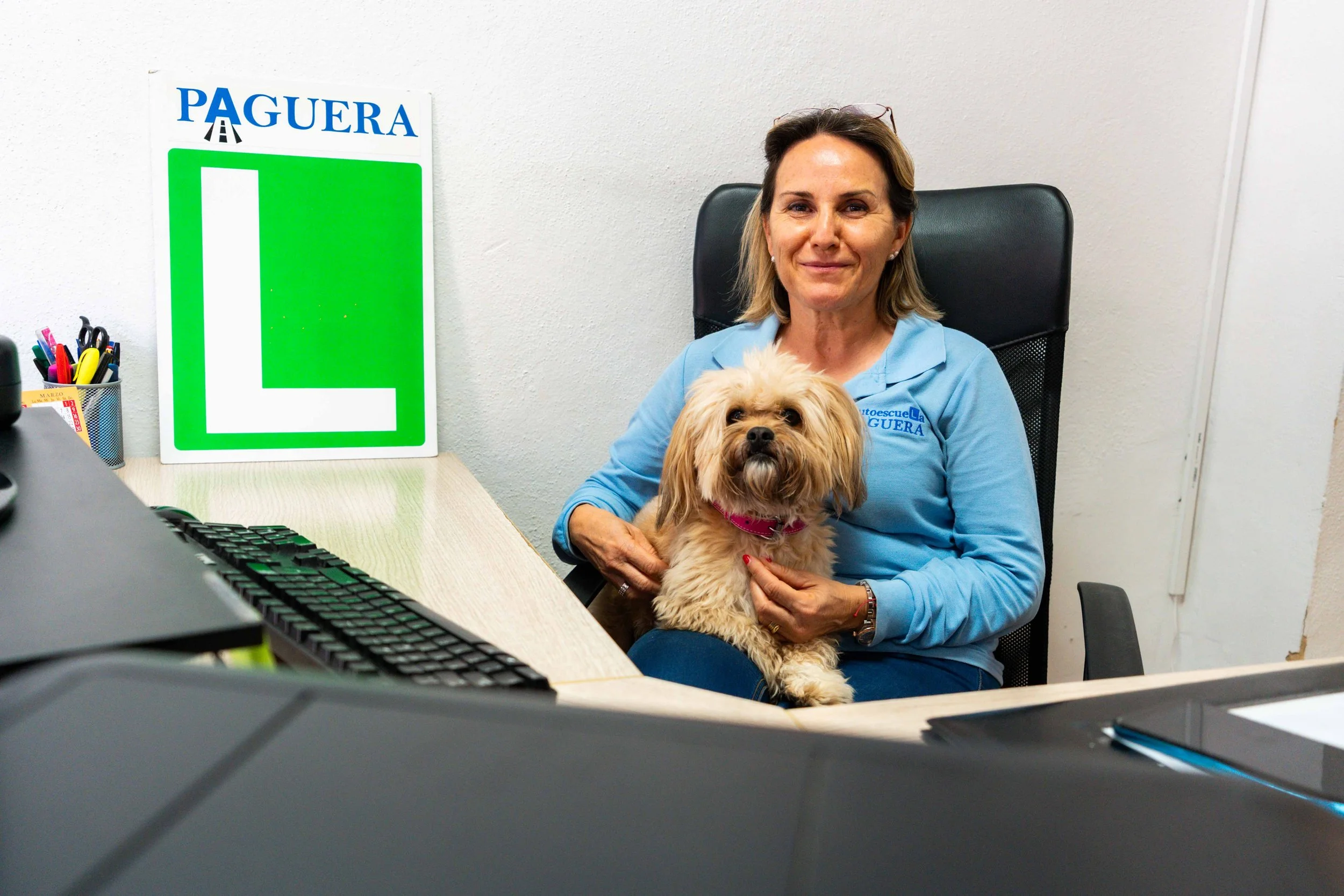 Una mujer sentada en una oficina, sosteniendo a un perro pequeño de color marrón, con fondo de pared blanca y cartel verde y blanco que dice 'Paguera'.
