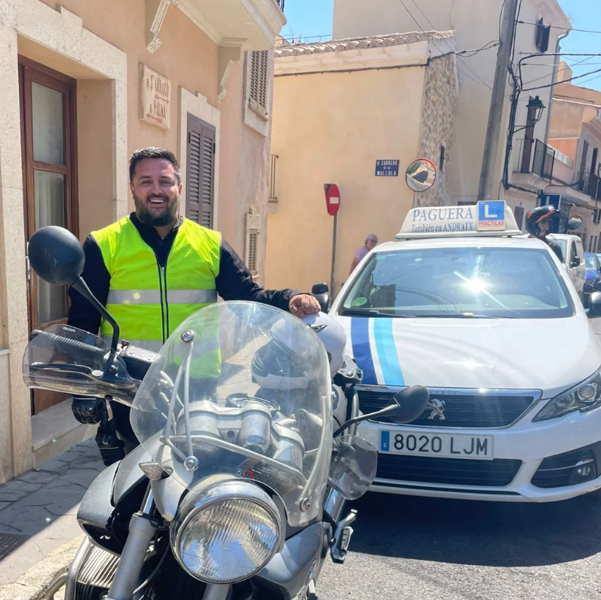 Un hombre con chaleco reflejante de pie junto a una motocicleta en una calle de un pueblo, con un coche de policía detrás. La escena es en un día soleado en un área urbana.