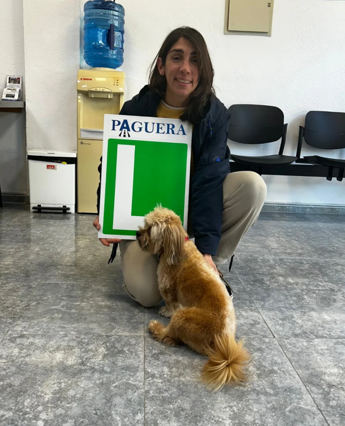 Mujer con un perro en una sala de espera, sosteniendo un cartel de 'Paguera' con un espacio en blanco en el centro.