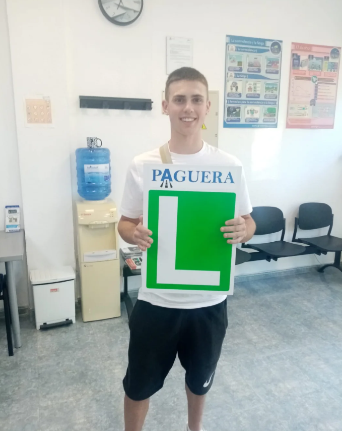 Joven sonriendo, sosteniendo cartel con letra L y la palabra 'Aguera', en sala de espera o oficina.