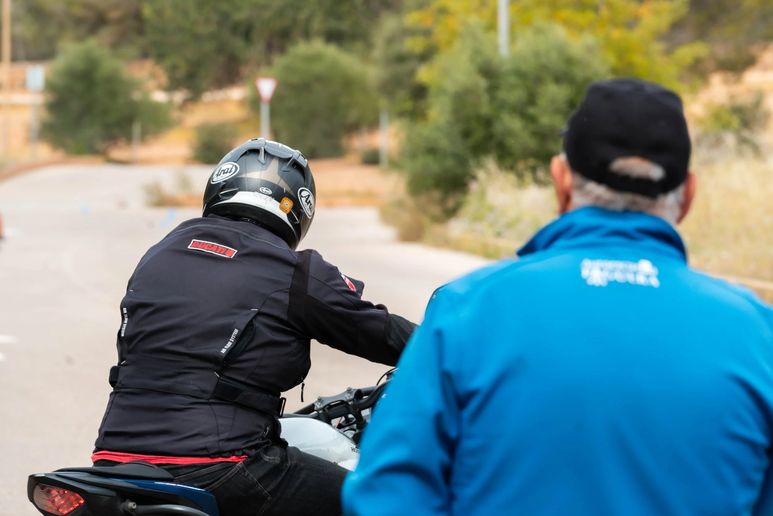 Dos personas en motocicleta en la carretera, una con casco y chamarra negra y otra con chamarra azul y gorra, con árboles y señal de tránsito en el fondo.