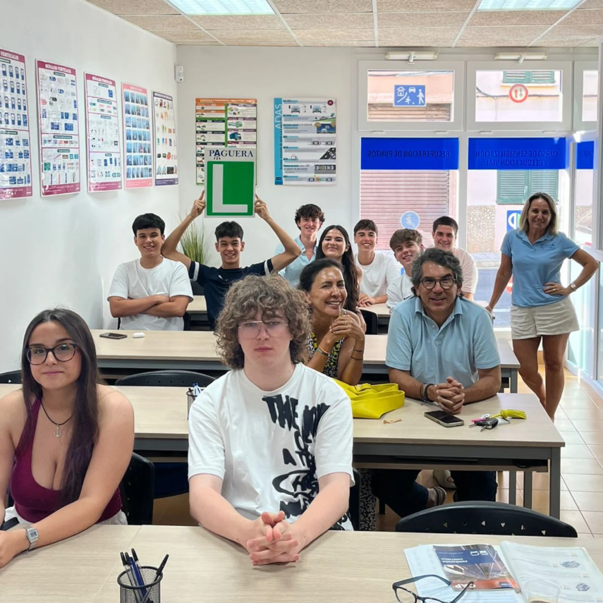 Grupo de estudiantes de autoescuela con instructor en salón de clases, algunos con señal de L de aprendizaje, posters y señalización en las paredes, y puertas con instrucciones de seguridad.