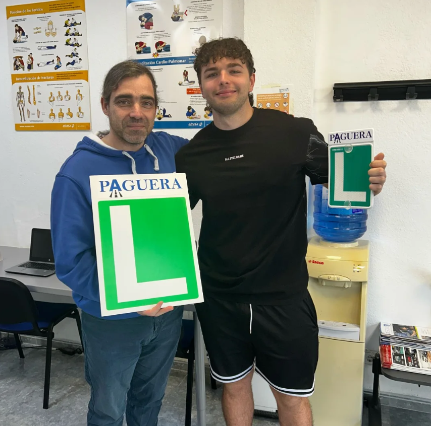Dos hombres sosteniendo carteles de la letra 'L' en letras grandes en un entorno de oficina o consulta médica, con posters en las paredes y una fuente de agua al fondo.