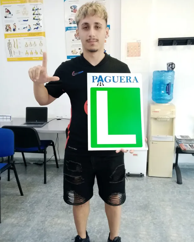 Joven sosteniendo un cartel que dice 'PAGUERA L', en una oficina o aula con mesas, sillas y una máquina dispensadora de agua.