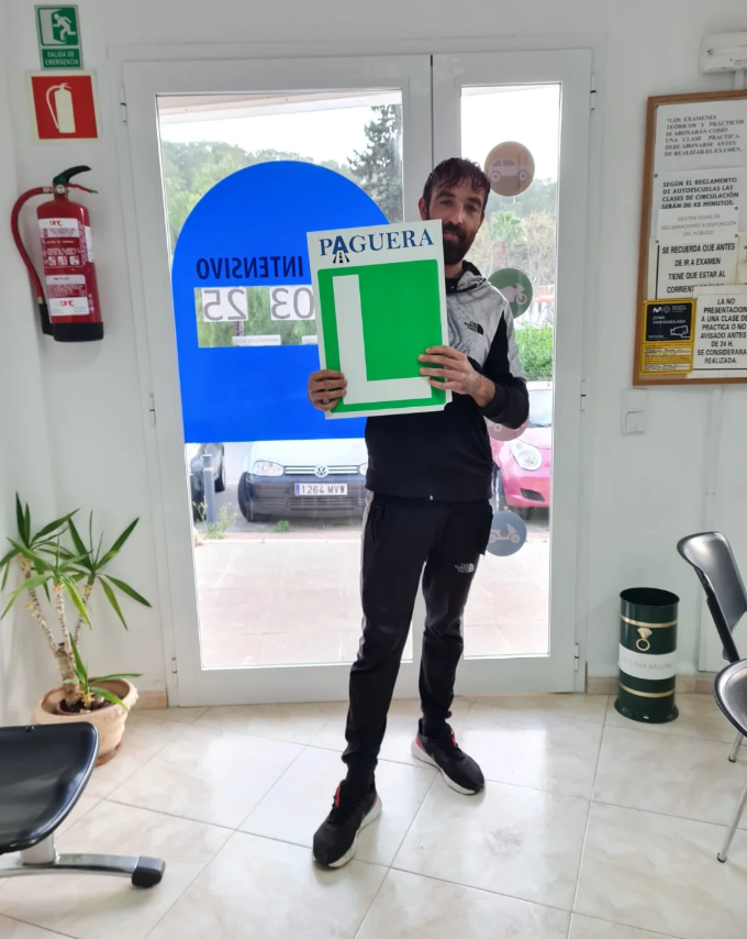 Hombre sosteniendo un cartel de fútbol en una zona de espera de un establecimiento, con puertas de vidrio y autos estacionados afuera.