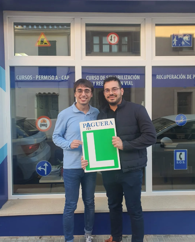 Dos hombres sonriendo frente a una ventana de una oficina de autos, uno de ellos sostiene un cartel verde con una 'L' blanca y la palabra 'PAGUERA'.