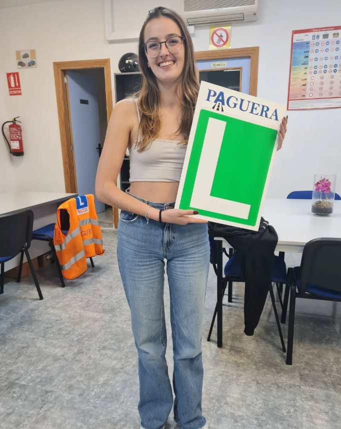 Joven mujer con gafas y ropa casual, sosteniendo un cartel que dice 'PAGUERA' y tiene una gran letra 'L', en un aula o sala de capacitación.