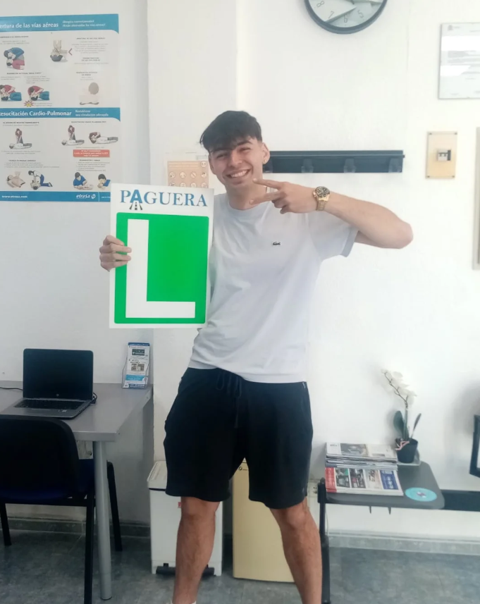 Joven sonriendo y señalando un cartel que dice 'PAGERA' con una letra 'L' grande, en un entorno de oficina o sala de espera.