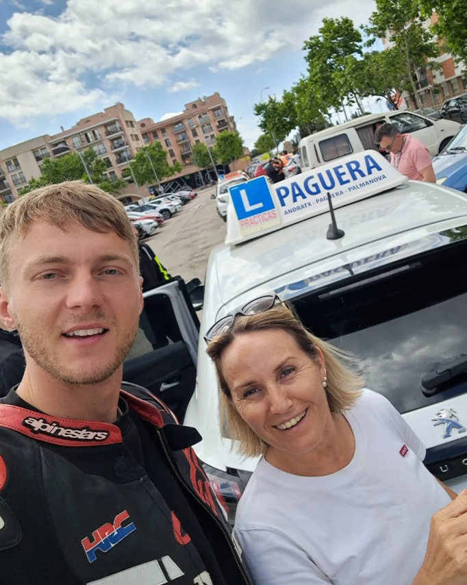 Dos personas en un auto de enseñanza de manejo en un estacionamiento, con un cartel que dice 'Paguera' en el techo del auto. Nubes y edificio de apartamentos de fondo.