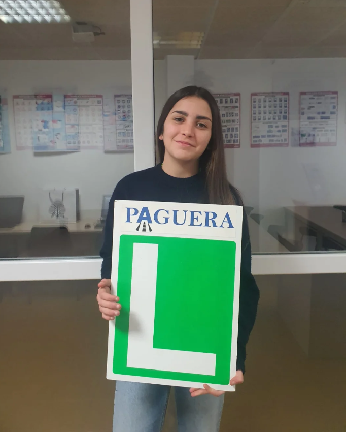 Mujer joven sosteniendo un cartel que dice "PAGUERA" arriba y tiene una letra roja grande "L" en verde.