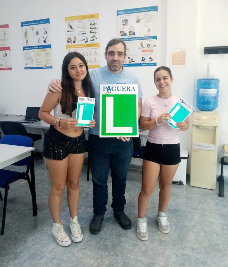 Tres personas en un aula, dos jóvenes y un adulto, sosteniendo señales con la letra 'L' y la palabra 'PAGUERA', en un ambiente de aprendizaje con carteles en la pared que parecen ser de salud o primeros auxilios.
