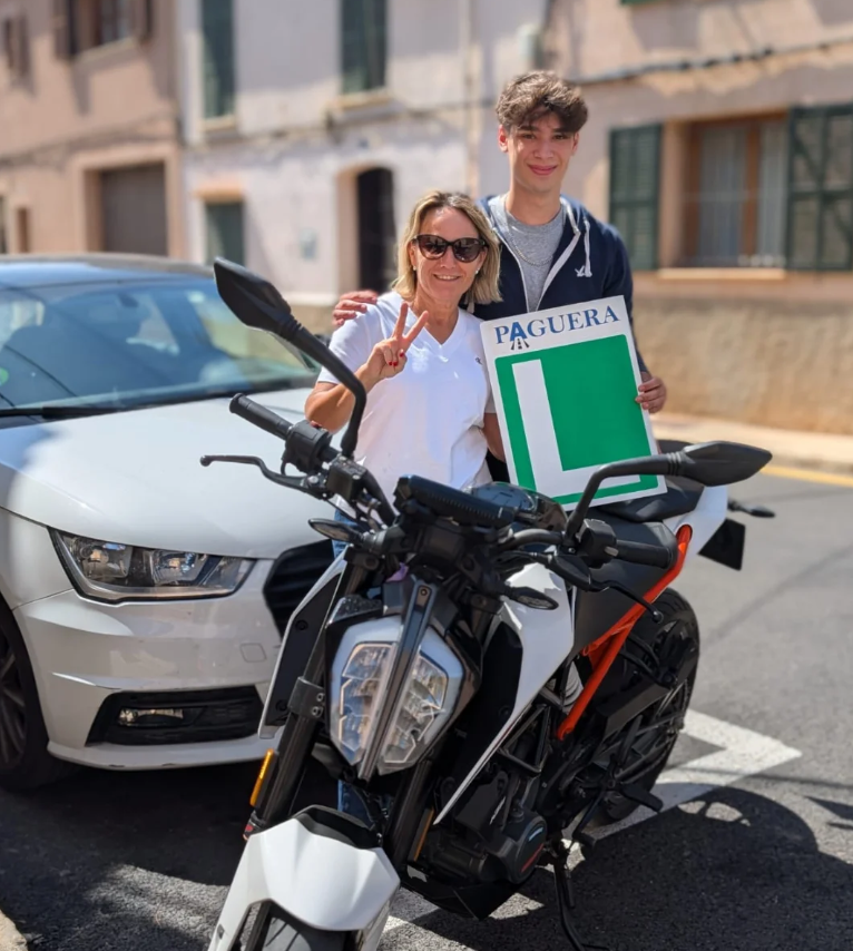 Una mujer y un joven posando juntos en una calle con un coche y motocicleta, la mujer hace señal de paz y sostienen un cartel que dice 'Paguera' y una letra 'L' de aprendiz de conducir.