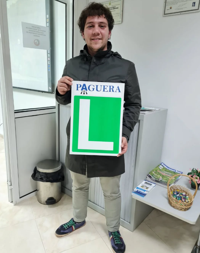 Joven sonriendo, sosteniendo señal de 'L' verde con la palabra 'Paguera' en un entorno interior, posiblemente una oficina o centro de atención.