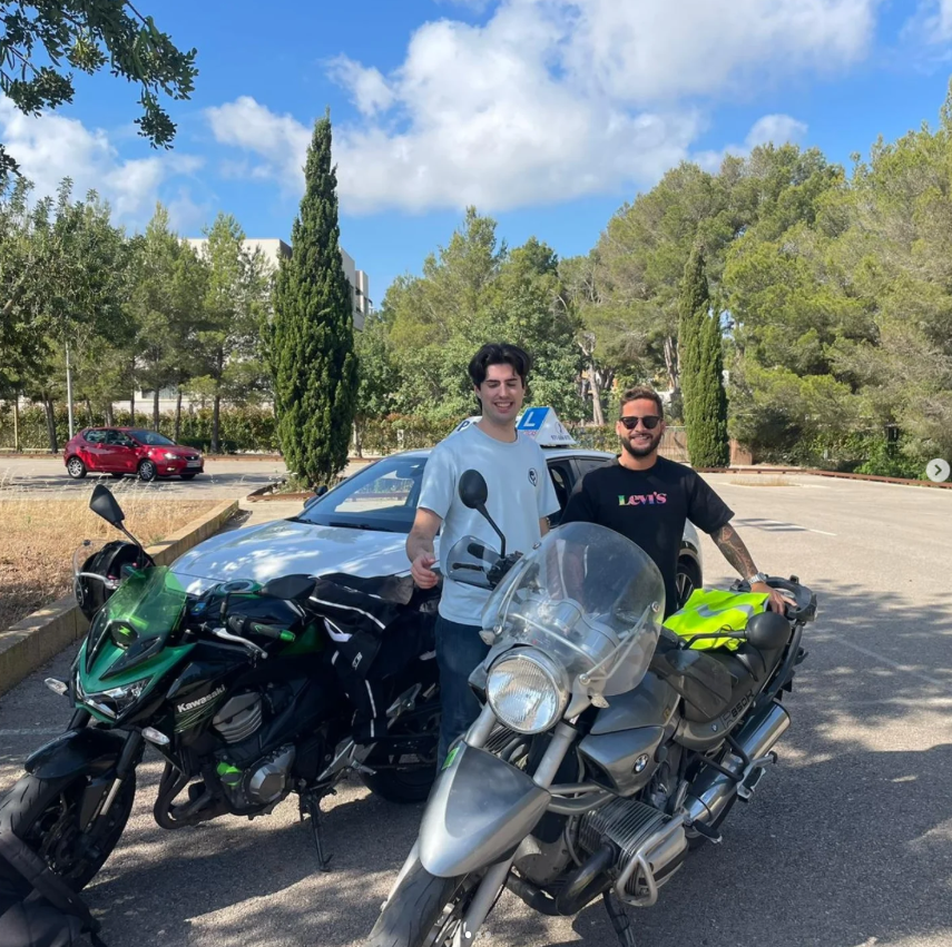 Dos hombres sonrientes junto a motocicletas en un estacionamiento rodeado de árboles y un coche azul, en un día soleado con cielo parcialmente nublado.