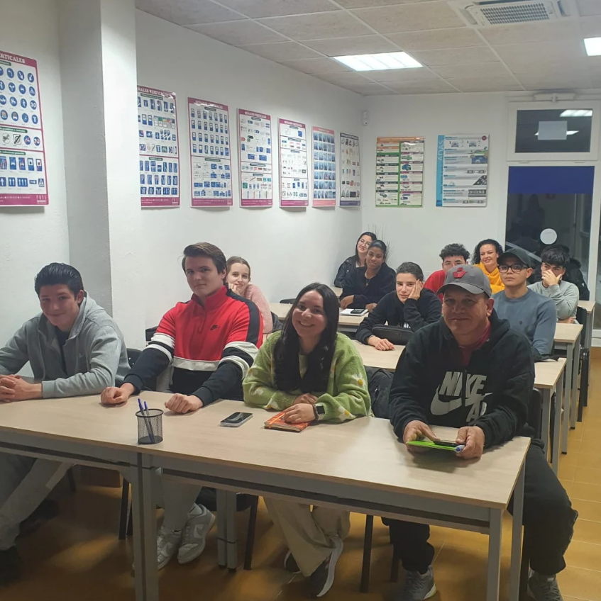 Grupo de estudiantes en un aula, sentados en mesas, mirando hacia la cámara, con posters instructivos en las paredes.