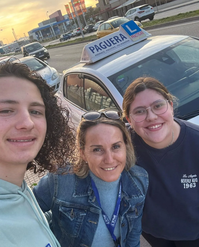 Tres personas posando para una selfie frente a un auto de autoescuela con señal de L y cartel de 'Paguera Practicas' en un día soleado, en una calle con otros autos y edificios en el fondo.