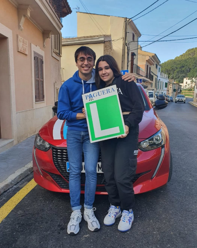 Jóvenes con diploma en la mano, frente a un coche rojo en una calle de pueblo, en un día soleado.