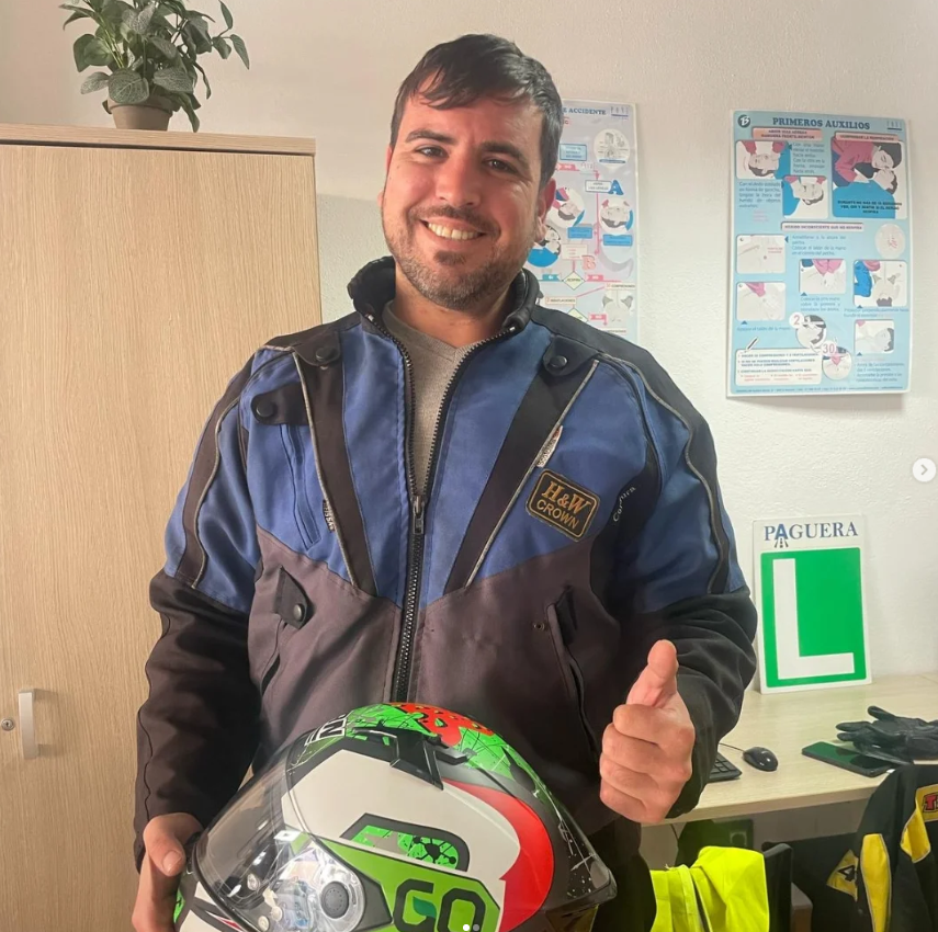 Hombre sonriente con chaqueta de motorista y casco, dando pulgar arriba en una estación de primeros auxilios, con plantas y carteles de instrucciones en el fondo.