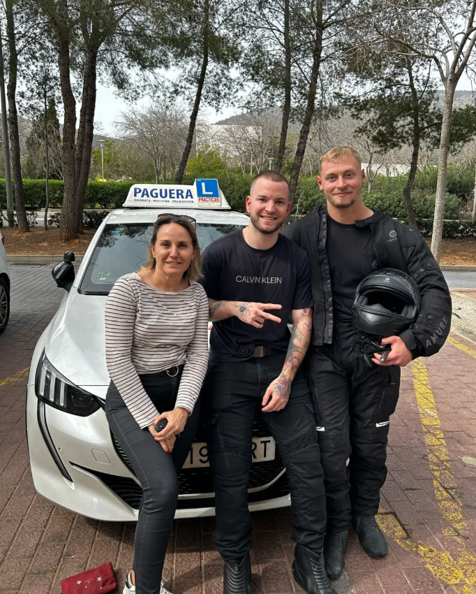 Tres personas posando frente a un coche, una mujer y dos hombres, en un entorno exterior con árboles y un estacionamiento.