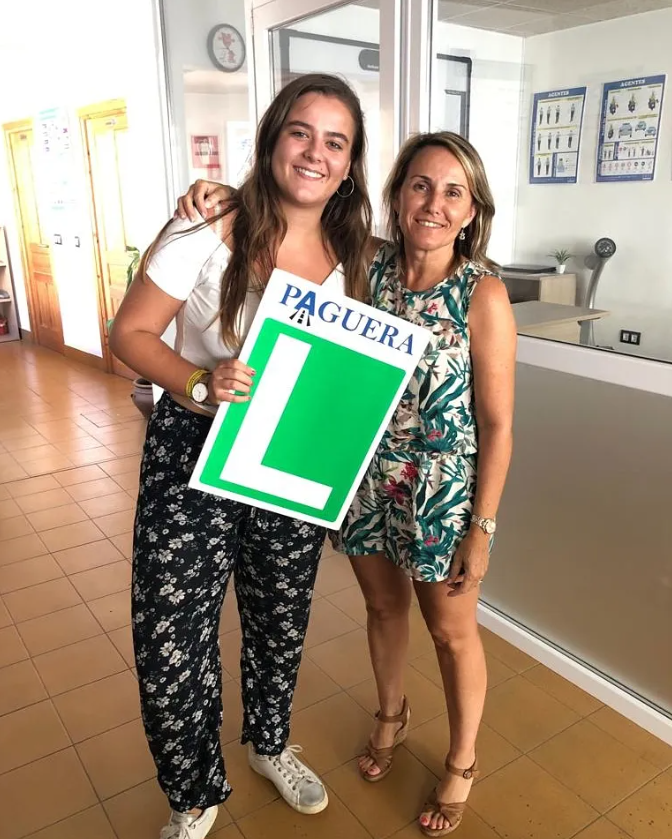 Dos mujeres sonriendo juntas en un gimnasio, sosteniendo un cartel que dice 'PAGUERA' y tiene un símbolo de una 'L' verde, indicando que una de ellas está recibiendo clases de conductor aprendiz.