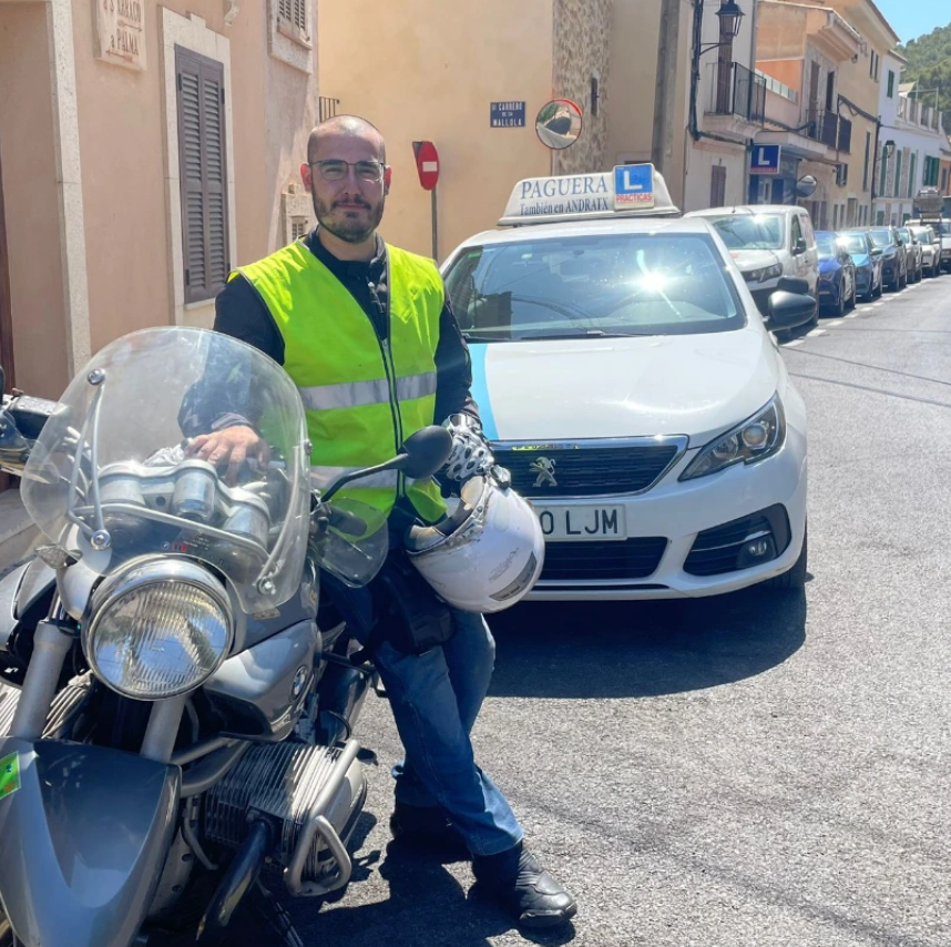 Un hombre con chaleco reflectante sobre motocicleta y casco blanco, en una calle con autos estacionados, frente a un coche blanco con señal de instructores de autoescuela y una señal de 'L' para conductores principiantes.