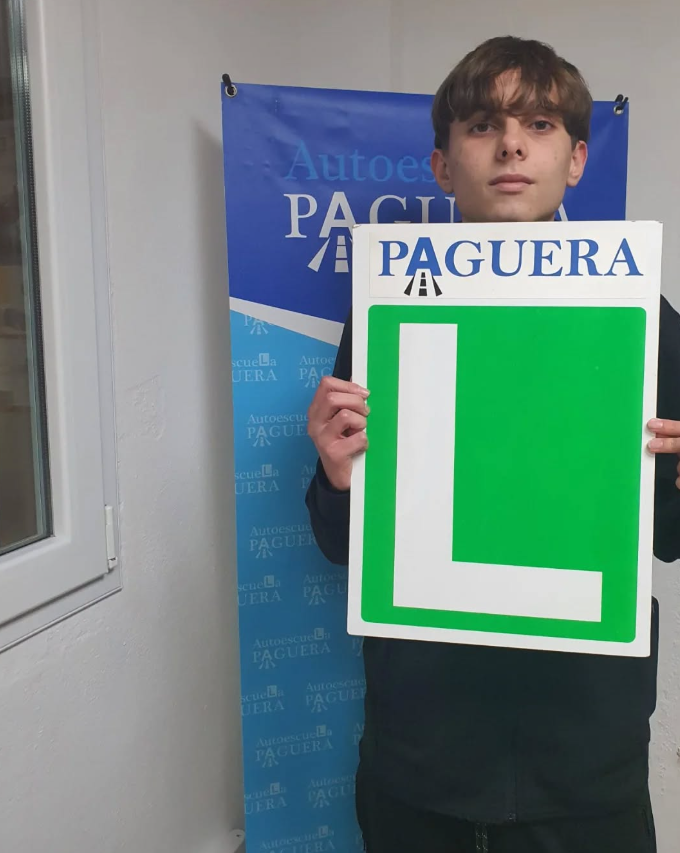 Joven sosteniendo cartel con letra 'L' grande, fondo con cartel azul que dice 'Autoscuela Paguera'.