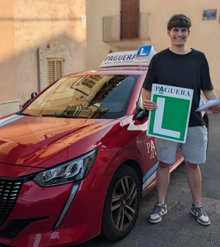 Joven sonriente sosteniendo un cartel con una 'L' grande, de pie junto a un coche rojo con señal de 'PAGUERA' en el techo, en una calle de un vecindario.