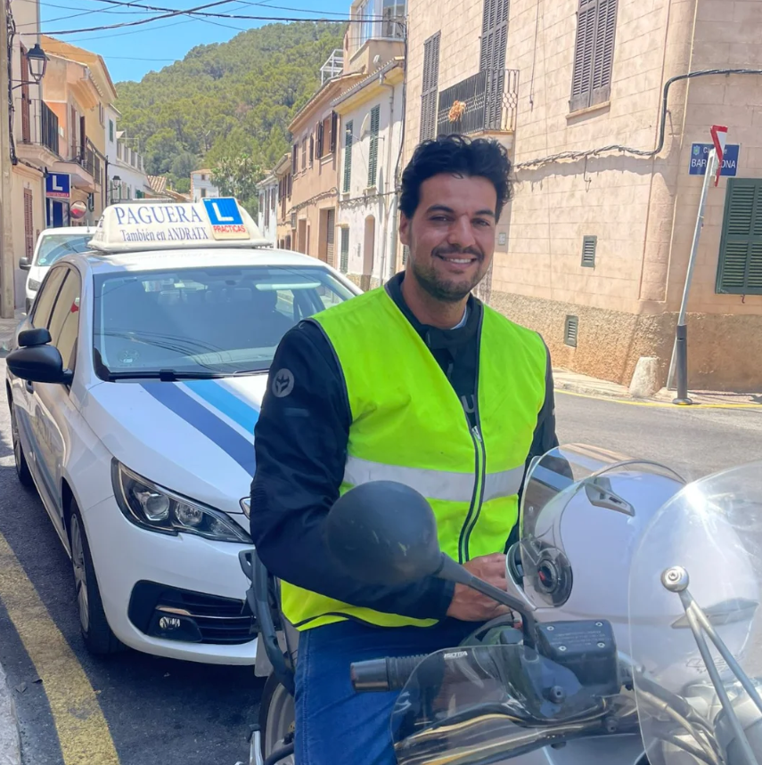 Un hombre con casco y chaleco reflectante verde sentado en una motocicleta en una calle asfaltada, con un coche policial al fondo, en un entorno urbano con edificios y colinas verdes.