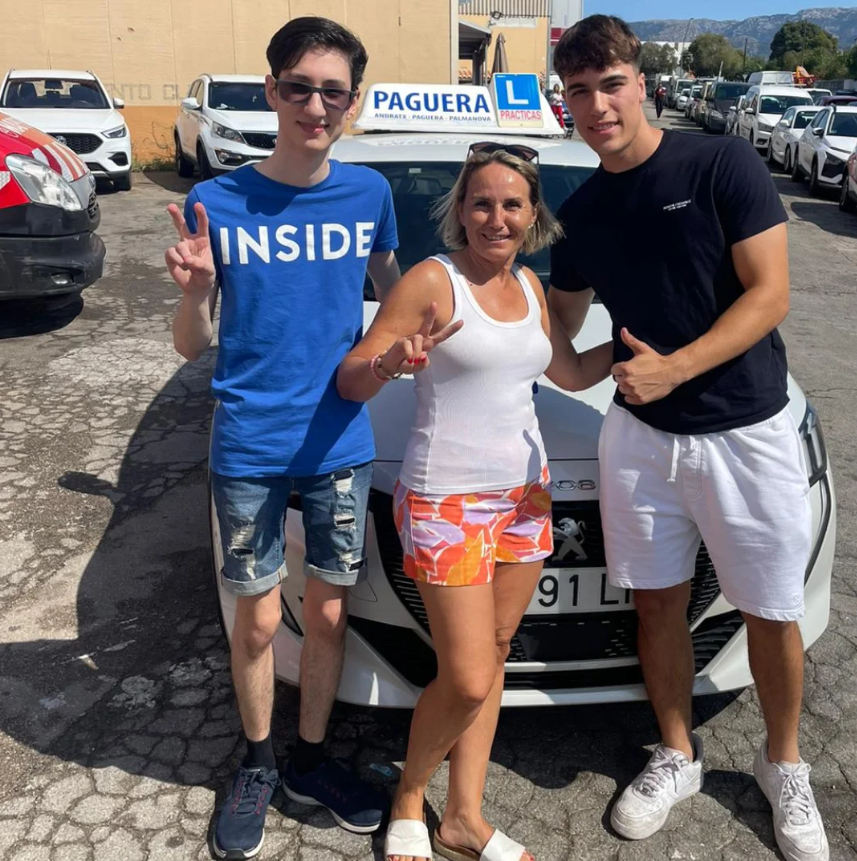 Tres personas posando en frente de un coche con un letrero de 'PAGUERA' en la azotea, en un estacionamiento, con autos y montañas al fondo, una mujer y dos jóvenes hombres.