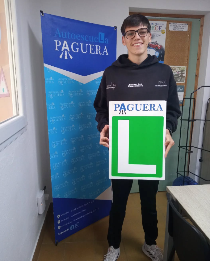 Joven sonriente sosteniendo un cartel con la letra 'L' en color verde y la palabra 'PAGUERA' en la parte superior, en un espacio cerrado con una pancarta azul que dice 'Autoescuela PAGUERA'