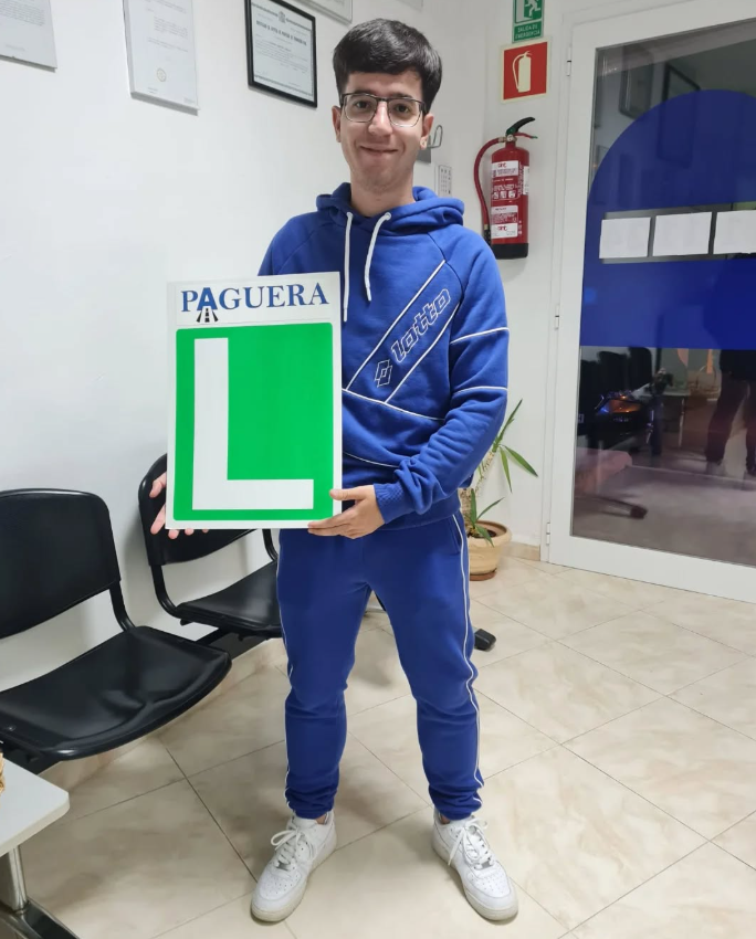Joven con gafas y camiseta azul, sosteniendo una señal de pezón y un cartel grande con una letra L, en una sala de espera.