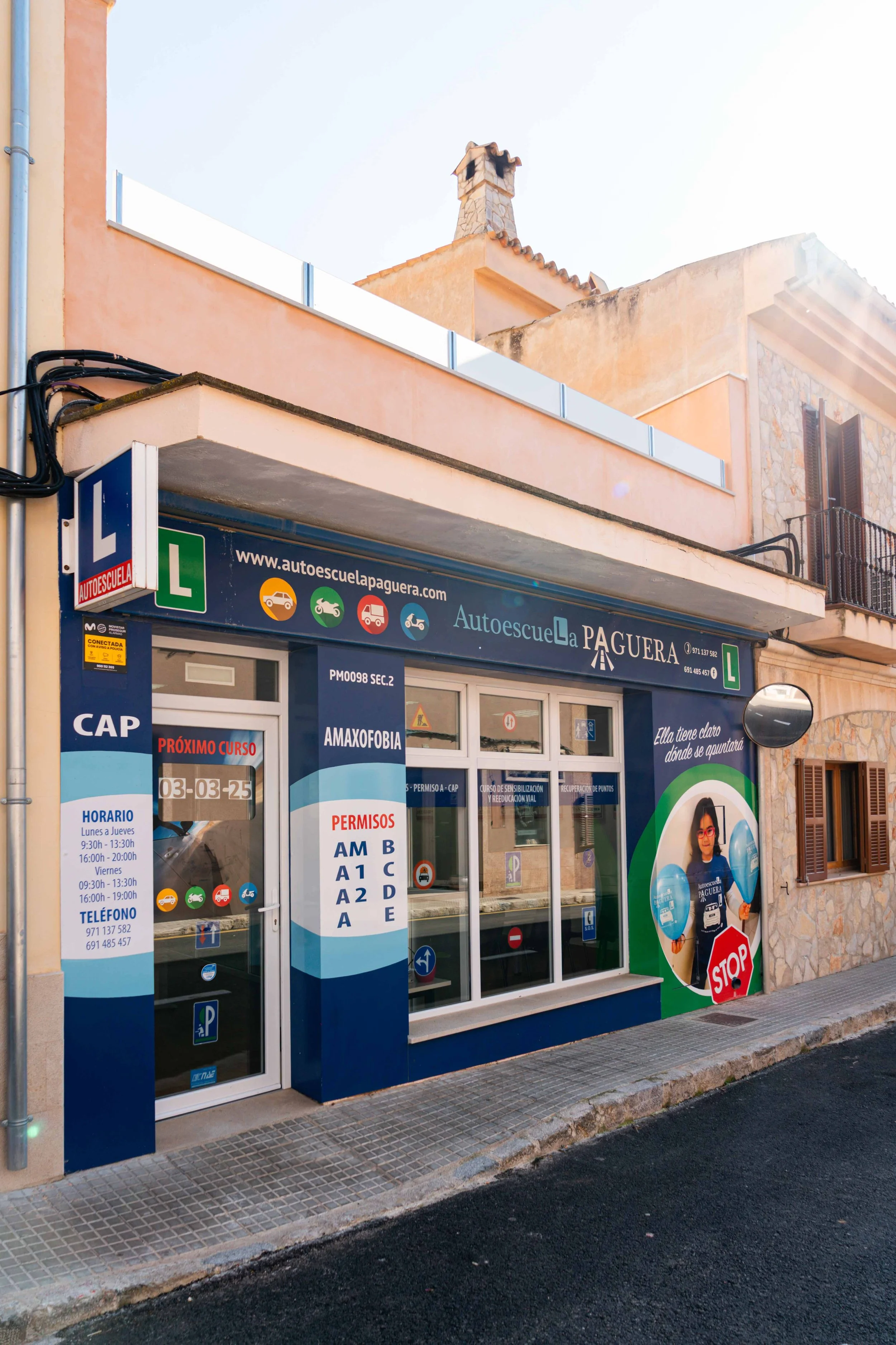 Fachada de una autoescuela llamada 'Autoescuela La Paguera' con cartelera sobre permisos de conducir y clases, y un cartel con la web www.autoescuelapaguera.com.