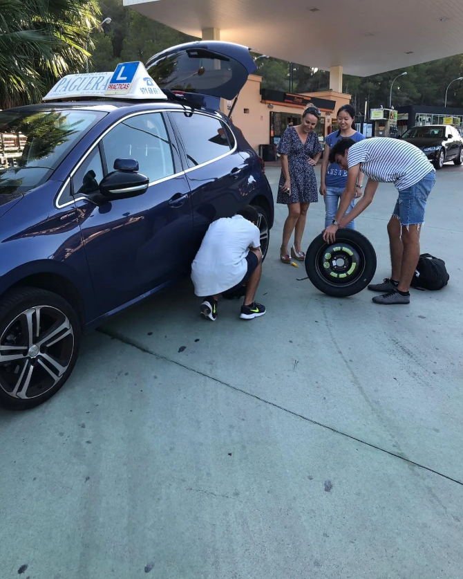 Grupo de personas revisando una rueda de coche en una estación de servicio, con un coche oscuro y un cartel de escuela de manejo en el techo.