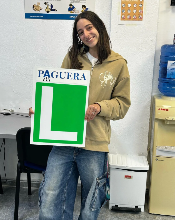 Joven sonriente sosteniendo un cartel verde y blanco con la palabra 'PAGUERA' y una 'L' grande, en un aula o oficina.