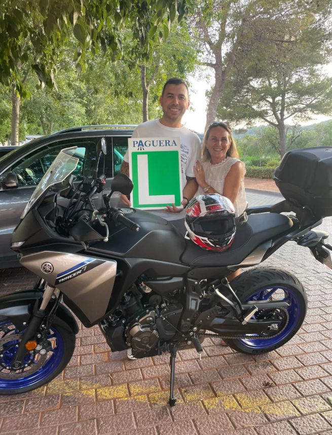 Hombre y mujer sonreíntes, sosteniendo un letrero con una 'L' y la palabra 'Paguera', detrás de una motocicleta negra y gris con casco protector sobre el asiento, en un área con árboles y autos estacionados.