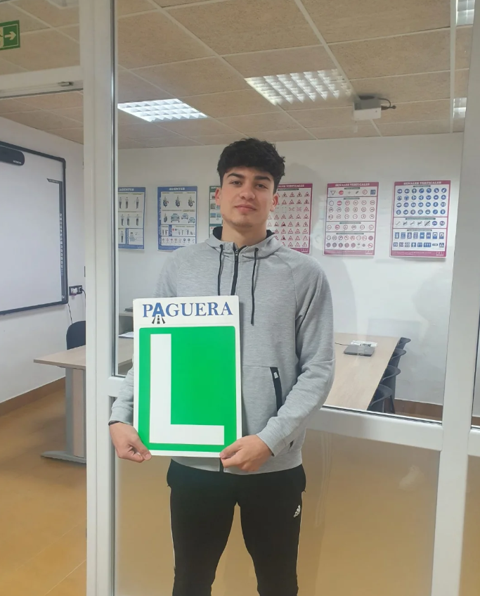 Joven con sudadera gris sostiene cartel con una 'L' grande en verde, señal de curso de aprendizaje de manejo, en aula con pósteres de señales de tránsito.
