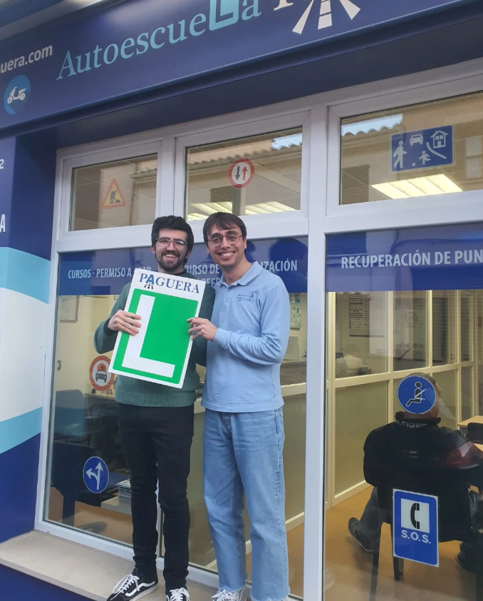 Dos hombres sonriendo y sosteniendo un cartel con una letra 'L' grande, fuera de una autoescuela llamada 'Autoescuela La'. Uno lleva gafas y una camiseta gris, el otro lleva gafas y una camisa azul. La autoescuela tiene ventanas con señalización de t