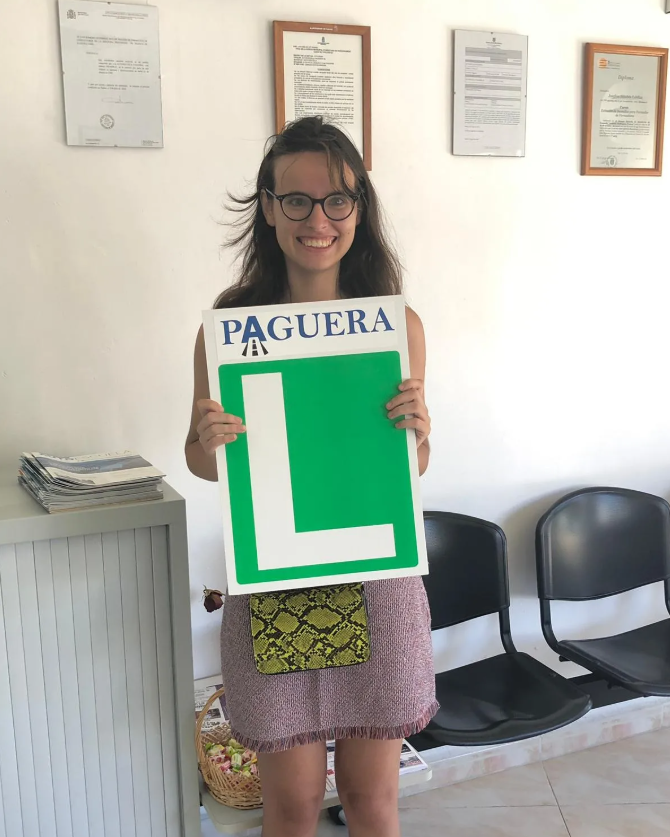 Joven mujer sonriendo, sosteniendo una señal de aprendizaje de manejo con la palabra 'PAGUERA' y una gran letra 'L' verde, en un consultorio o oficina.