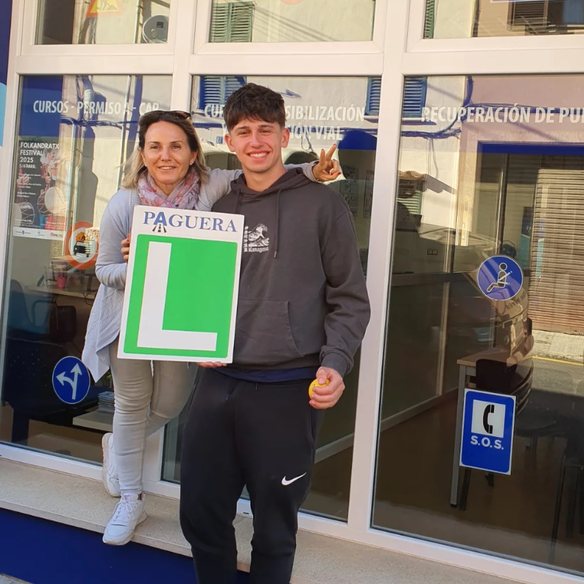 Joven con gafas de sol sosteniendo un cartel con una 'L' verde y la palabra 'PAGUER', acompañado de una mujer que hace una señal de paz. Están frente a una ventana de un centro de aprendizaje con señalización de clases de recuperación y permisos de c
