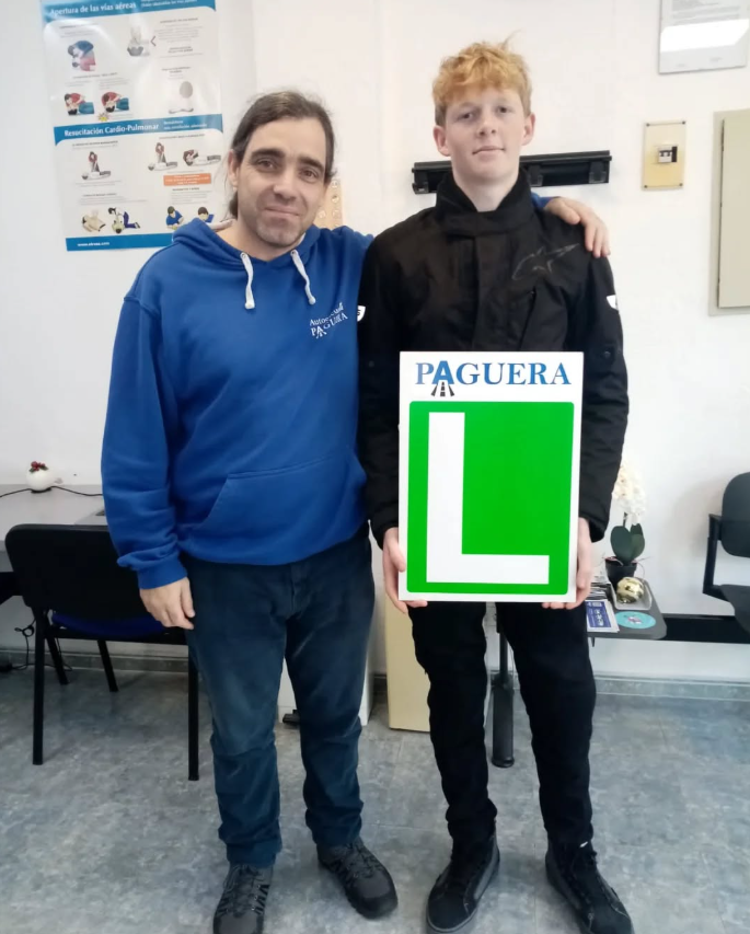 Persona con placa de 'L' para conductores principiantes, acompañada por un instructor, en un entorno de sala de aprendizaje o examen de manejo.