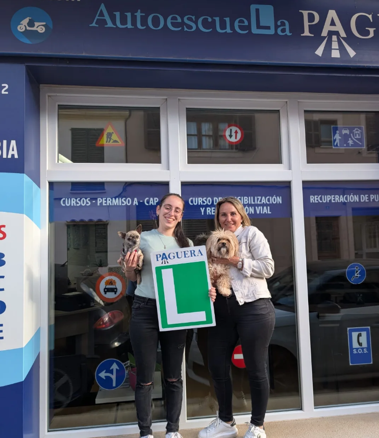 Dos mujeres sosteniendo perros pequeños frente a una oficina de autoescuela llamada 'Autoescuela La PAG', una de ellas con un cartel de 'L' de aprendizaje vocacional.