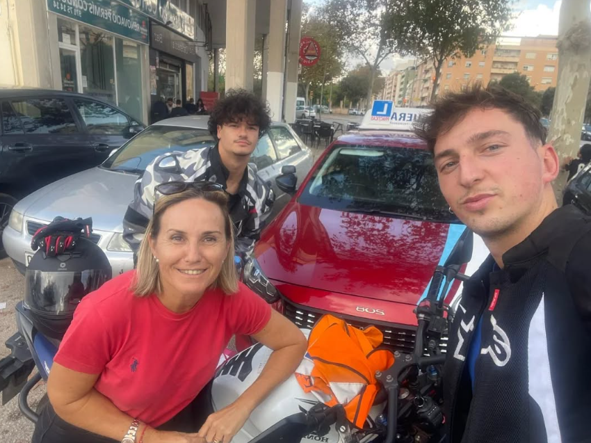 Grupo de tres personas, dos hombres y una mujer, posando en la calle frente a automóviles y motocicleta, con árboles y edificios en el fondo.