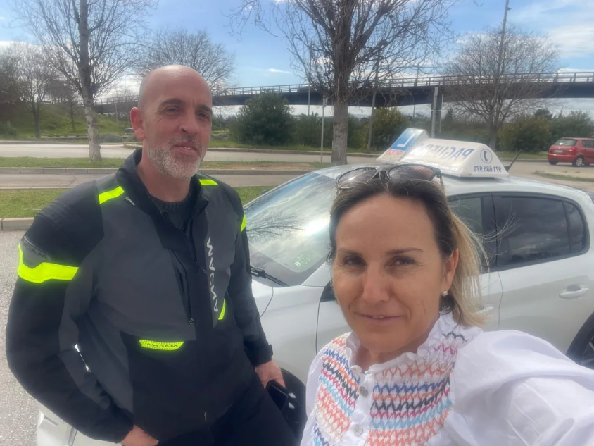 Dos personas, un hombre con chaqueta de motociclista y una mujer con blusa blanca, tomando una selfie frente a un auto de taxi en un día soleado.