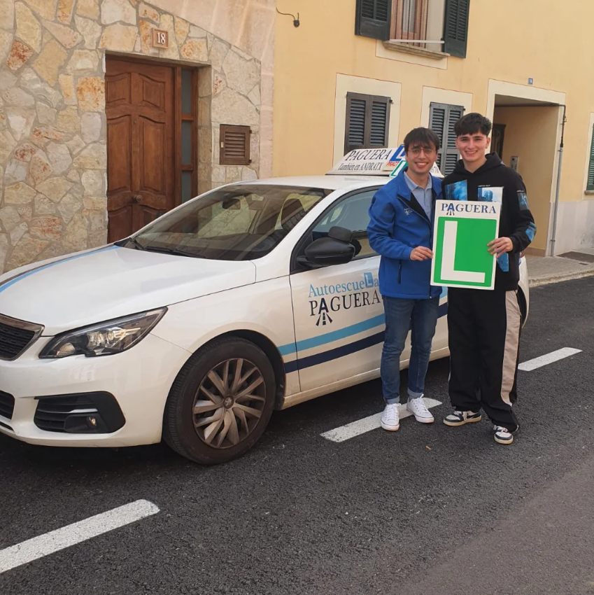 Dos jóvenes sosteniendo un cartel de 'L' para autoescuela, junto a un coche blanco con logotipo de la autoescuela 'Paguera' en una calle con casas de paredes de piedra y ventanas con contraventanas.