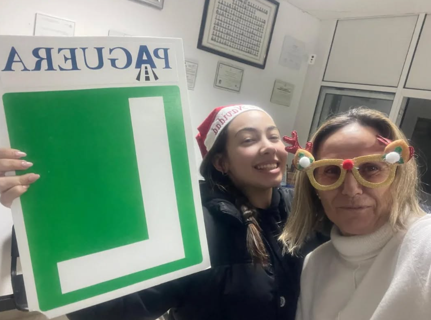 Dos mujeres en una oficina, una con un gorro de Navidad y la otra con gafas de fiesta, tomando una selfie. La mujer con gorro sostiene un cartel grande con texto invertido que dice 'PAGUEBLA'.