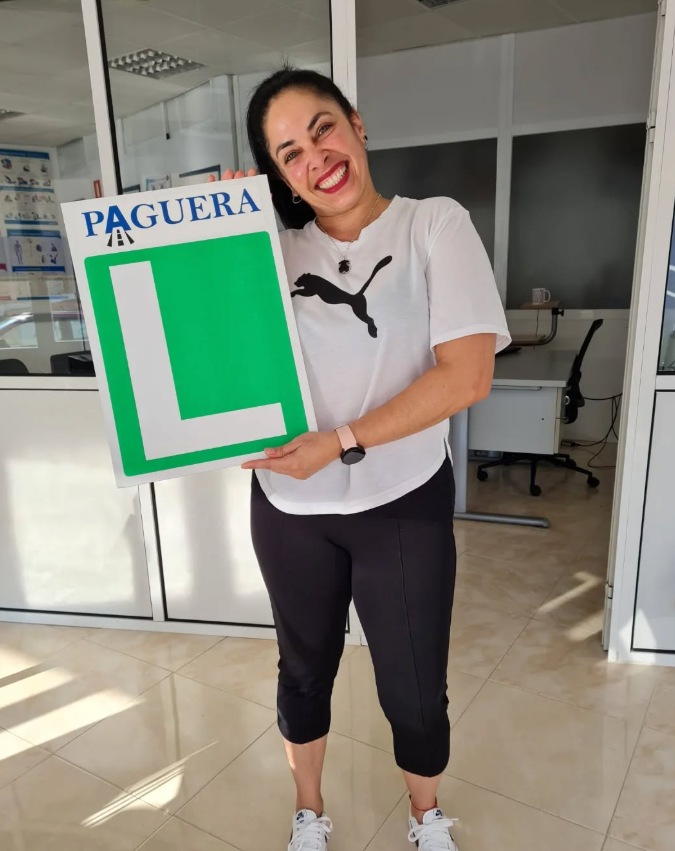 Mujer sonriendo sosteniendo un cartel grande con la letra 'L' en color verde y la palabra 'PAGUERA' en la parte superior, en una oficina.