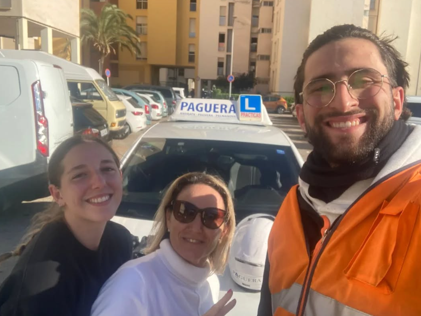 Tres personas posando frente a un coche de autoescuela con señal de 'PAGUERA' y una 'L' azul, en un estacionamiento exterior, con edificios y palmeras al fondo.