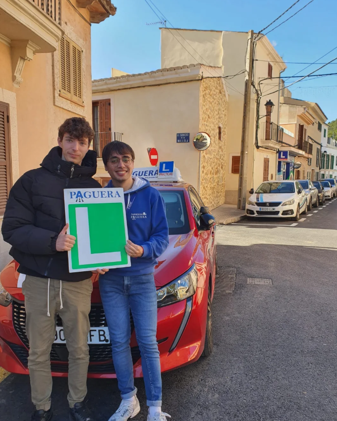 Dos jóvenes conductores posan frente a un coche rojo de autoescuela, uno sostiene un cartel verde con una L yotro con un cartel que dice 'PAGUERA'.