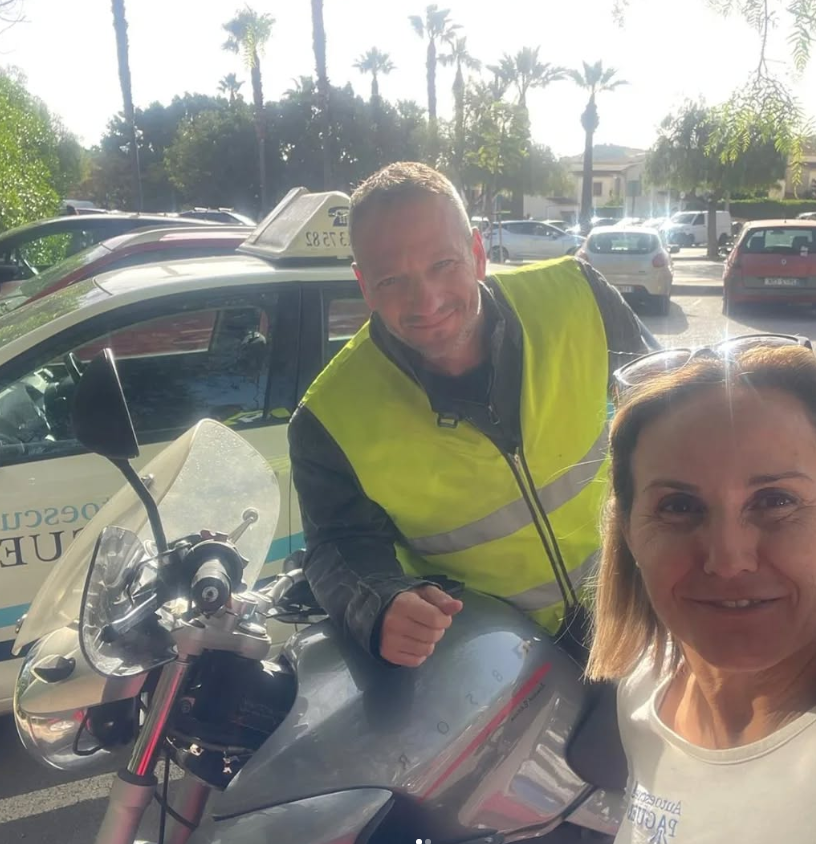 Una mujer y un hombre sonriendo en una estación de policía, con autos estacionados y palmeras en el fondo durante el día.